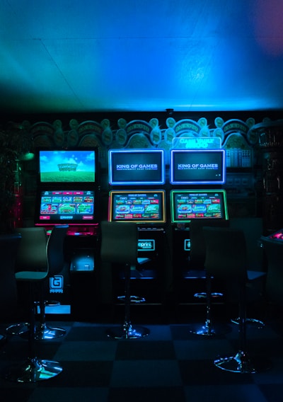 Dimly lit gaming arcade featuring a variety of brightly illuminated slot machines and video gaming screens. Neon signs and colorful displays create a vibrant atmosphere. The arrangement of gaming machines suggests an inviting space for entertainment, with chairs positioned for players.